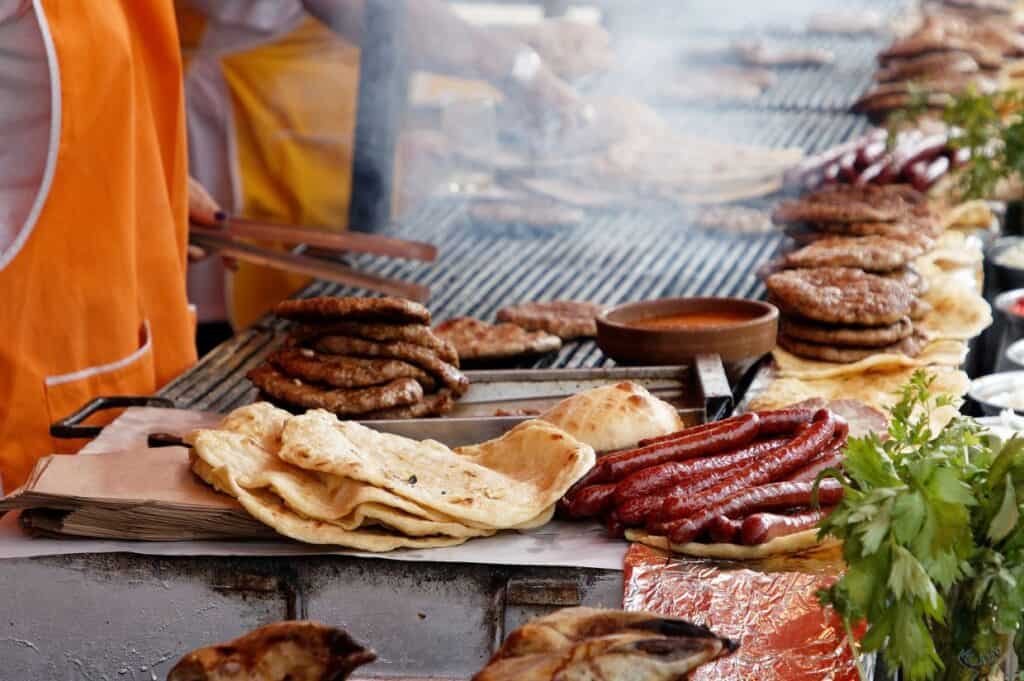 A variety of grilled meats, flatbreads, and sausages are being cooked on an outdoor grill at a lively bbq festival, with chefs in orange aprons working in the background.
