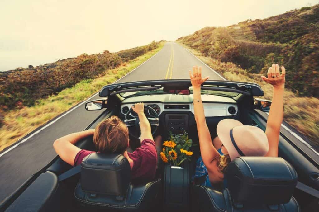 Two people ride in a convertible on an empty road, with one raising their hands. Flowers are on the console, and the scene is sunny with open countryside.