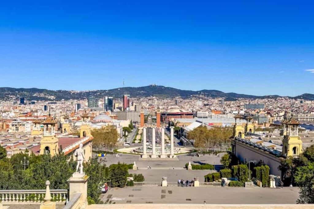 A panoramic view of Barcelona featuring four columns, buildings, and mountains in the background under a clear blue sky.